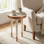 Aydınlık bir odada, krem rengi puf dokulu bir halının üzerinde duran, beyaz ve doğal ahşap detaylı yuvarlak yan sehpa. Sehpanın üst yüzeyinde kabartmalı sarmal desenler bulunur; üzerinde bir kupa kahve, üst üste dizili kitaplar ve küçük bir sukulent saksısı yer alıyor. Arka planda bej bir koltuk ve tül perdeli bir pencere görülüyor.