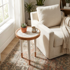 Aydınlık bir odada, krem rengi puf dokulu bir halının üzerinde duran, beyaz ve doğal ahşap detaylı yuvarlak yan sehpa. Sehpanın üst yüzeyinde kabartmalı sarmal desenler bulunur; üzerinde bir kupa kahve, üst üste dizili kitaplar ve küçük bir sukulent saksısı yer alıyor. Arka planda bej bir koltuk ve tül perdeli bir pencere görülüyor.