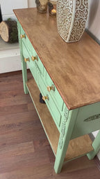 Mint yeşili boyalı, oyma detaylı çekmecelere sahip, üst kısmı ahşap kaplamalı dekoratif dresuar; üzerinde seramik vazo ve mumluklar, alt rafında dekoratif objeler bulunan vintage tarz konsol.